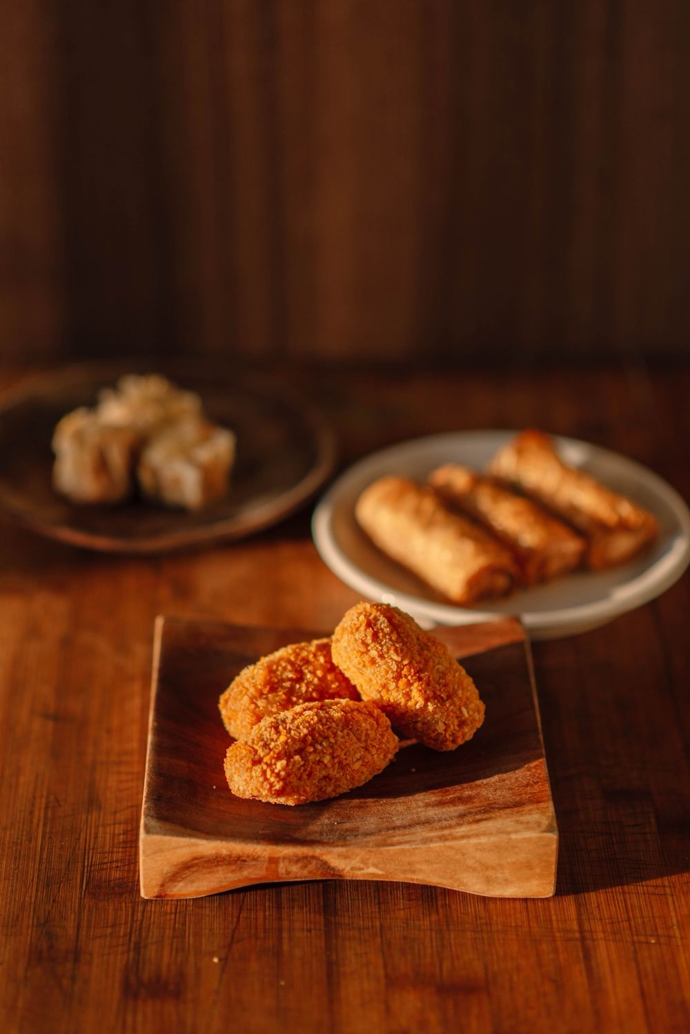 Krokante snacks op een houten plankje, met loempia's en siu mai op de achtergrond